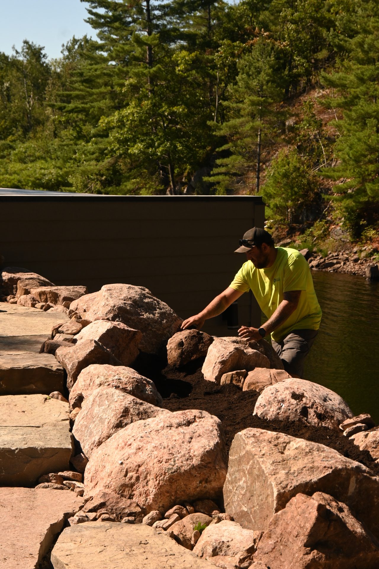 Man landscaping with rocks near water and trees.
