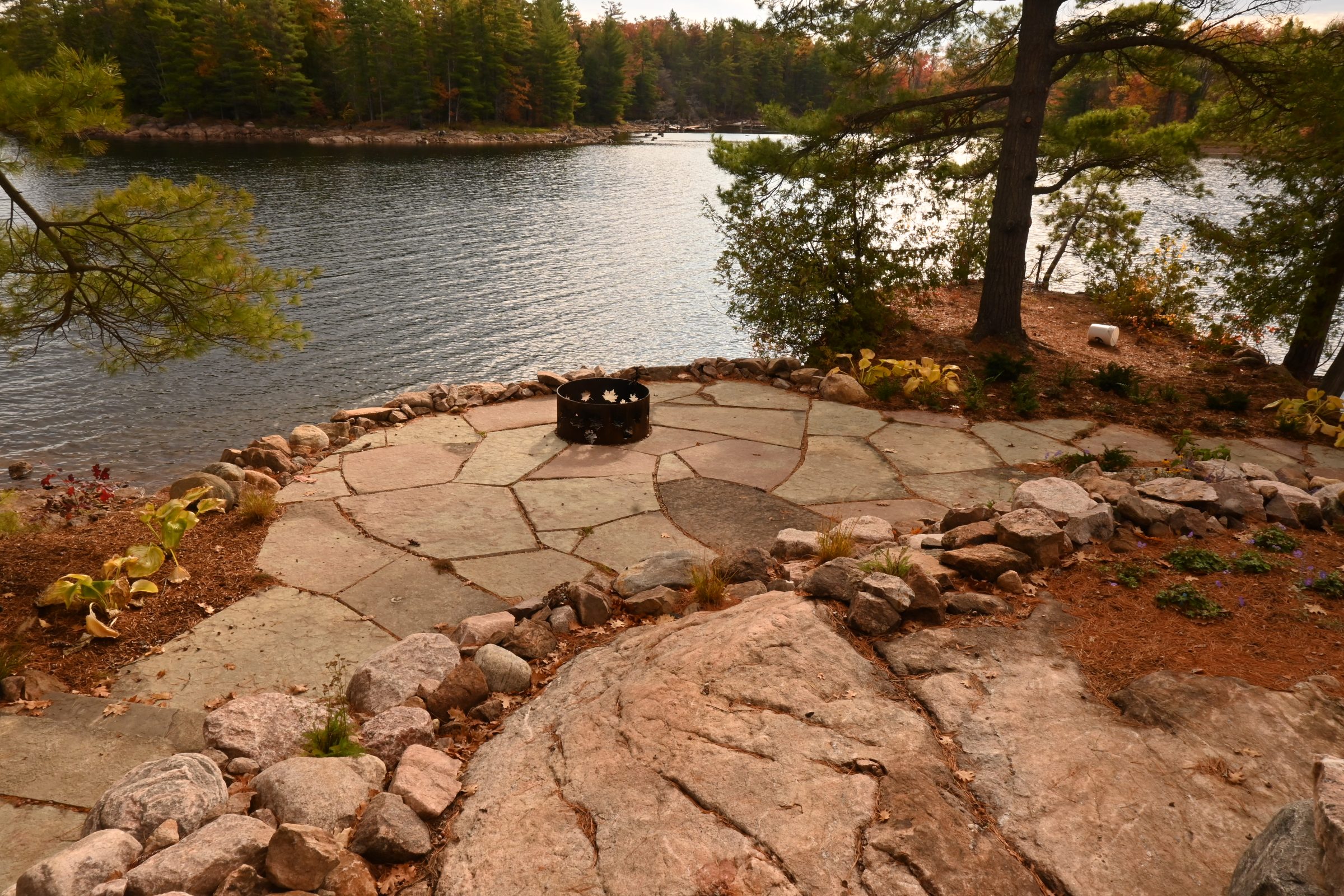 Lakeside stone patio with fire pit and trees.